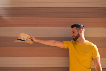 A handsome young man with blue eyes and a beard and a yellow T-shirt stretches out his arm with a hat in his hand. In the background a wall in shades of brown. The man smiles happily.