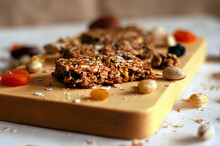 Bar With Flakes, Nuts And Dried Fruits On Light Wooden Board With Ingredients Scattered Side By Side On Gray Background. Board Is Located Diagonally. Natural Light Source, Selective Focus, Side View