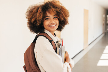 Wall Mural - Smiling girl standing in college corridor holding textbooks and looking at camera