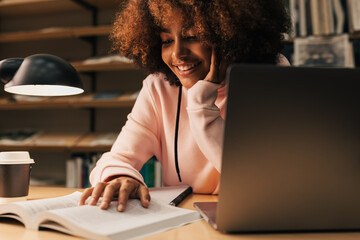 Wall Mural - Smiling student studying late at night. Young female using book and laptop at library desk.