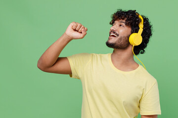 Wall Mural - Young singer happy Indian man 20s in basic yellow t-shirt headphones listen to music sing song in microphone isolated on plain pastel light green background studio portrait People lifestyle concept.