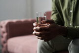 Fototapeta Łazienka - Hands of young troubled African American man holding glass of water while sitting on couch in office of his psychoalyst or psychologist