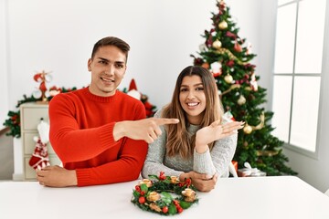 Canvas Print - Young hispanic couple sitting at the table on christmas amazed and smiling to the camera while presenting with hand and pointing with finger.