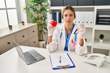 Canvas Print - Young blonde woman wearing doctor uniform holding blood and heart relaxed with serious expression on face. simple and natural looking at the camera.