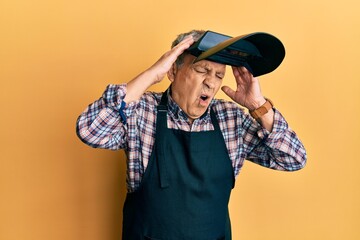 Wall Mural - Handsome senior man with grey hair wearing welding protection mask suffering from headache desperate and stressed because pain and migraine. hands on head.