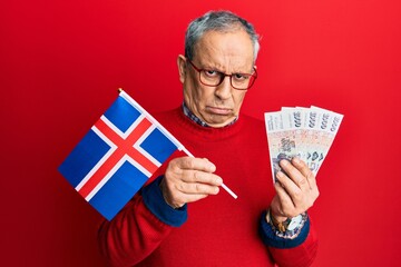 Canvas Print - Handsome senior man with grey hair holding iceland flag and icelandic krona banknotes skeptic and nervous, frowning upset because of problem. negative person.