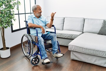 Canvas Print - Handsome senior man sitting on wheelchair at the living room smiling with happy face looking and pointing to the side with thumb up.