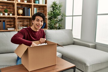 Sticker - Young hispanic man unboxing cardboard box sitting on the sofa at home.