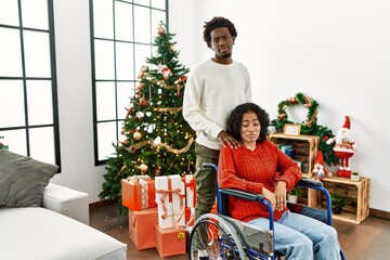 Poster - Young interracial couple with woman sitting on wheelchair by christmas tree depressed and worry for distress, crying angry and afraid. sad expression.