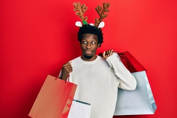 Poster - Young african american man wearing deer christmas hat holding shopping bags smiling looking to the side and staring away thinking.