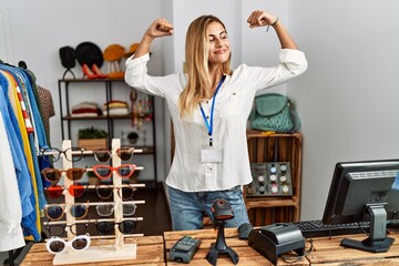 Poster - Blonde beautiful young woman working as manager at retail boutique showing arms muscles smiling proud. fitness concept.