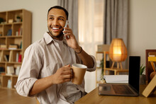 Positive Young African American Man With Coffee Talking On Cellphone, Using Laptop Computer At Home Office, Copy Space