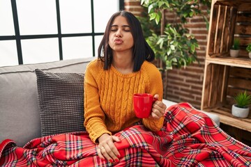 Poster - Young hispanic woman sitting on the sofa drinking a coffee at home looking at the camera blowing a kiss on air being lovely and sexy. love expression.