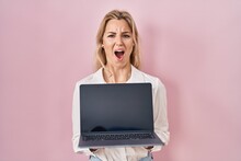 Young Caucasian Woman Holding Laptop Showing Screen Angry And Mad Screaming Frustrated And Furious, Shouting With Anger. Rage And Aggressive Concept.