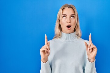 Poster - Young caucasian woman standing over blue background amazed and surprised looking up and pointing with fingers and raised arms.