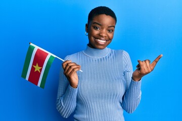 Poster - Young african american woman holding suriname flag smiling happy pointing with hand and finger to the side