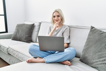 Sticker - Young caucasian woman using laptop at home sitting on the sofa looking confident at the camera smiling with crossed arms and hand raised on chin. thinking positive.