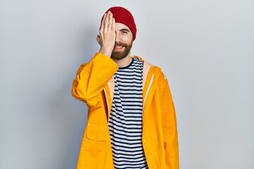 Wall Mural - Caucasian man with beard wearing yellow raincoat covering one eye with hand, confident smile on face and surprise emotion.