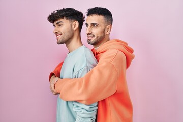 Sticker - Young hispanic gay couple standing over pink background looking to side, relax profile pose with natural face and confident smile.