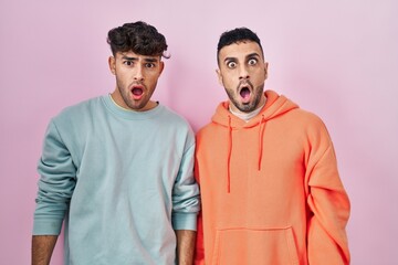 Young hispanic gay couple standing over pink background in shock face, looking skeptical and sarcastic, surprised with open mouth