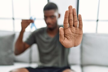 Poster - Young african american man holding blue ribbon sitting on the sofa at home with open hand doing stop sign with serious and confident expression, defense gesture