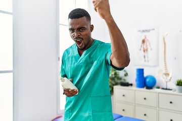 Wall Mural - Young african physiotherapist man holding massage body lotion annoyed and frustrated shouting with anger, yelling crazy with anger and hand raised