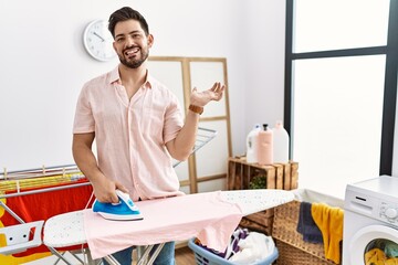 Poster - Young man with beard ironing clothes at home smiling cheerful presenting and pointing with palm of hand looking at the camera.