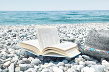 Open Book And Male Straw Hat On The Pebble Beach. Concept Of Reading And Relaxing In Summer Vacation. Beach Literature.