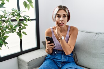 Sticker - Young beautiful woman using smartphone typing message sitting on the sofa hand on mouth telling secret rumor, whispering malicious talk conversation