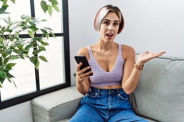 Sticker - Young beautiful woman using smartphone typing message sitting on the sofa pointing aside with hands open palms showing copy space, presenting advertisement smiling excited happy