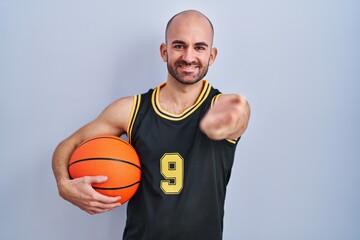 Sticker - Young bald man with beard wearing basketball uniform holding ball smiling cheerful offering palm hand giving assistance and acceptance.