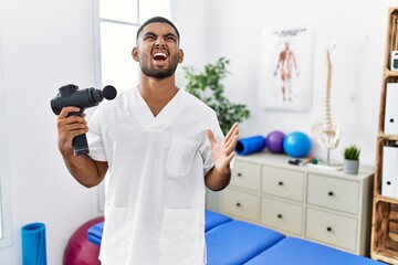 Canvas Print - Young indian physiotherapist holding therapy massage gun at wellness center crazy and mad shouting and yelling with aggressive expression and arms raised. frustration concept.