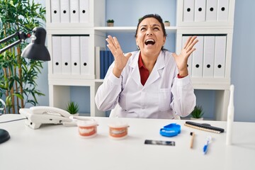 Sticker - Hispanic mature dentist woman working at the clinic crazy and mad shouting and yelling with aggressive expression and arms raised. frustration concept.