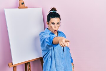 Poster - Young hispanic woman standing by painter easel stand pointing with finger surprised ahead, open mouth amazed expression, something on the front