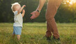 First baby steps. A young father helps a baby boy walk on the grass in the park. Parental care and support