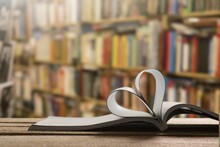 An Open Book Forming A Heart With Two Pages In The Center, In The Library. In The Background Are Shelves Full Of Books.