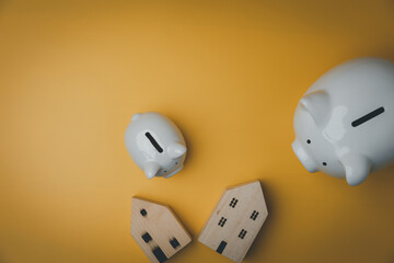 Flat lay, top view. Two white piggy banks with a small wooden house model on yellow background with copy space. Accounting, saving money, real estate investment, banking concept.