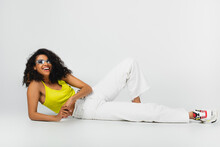 Happy African American Woman In Blue Sunglasses And Stylish Outfit Posing On Grey.