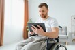 © Serhii - Man in wheelchair using tablet computer at home. Positive retired male with physical disability browsing web on touch pad, watching video online indoors.