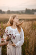 A young girl with long blond hair in a long white nightgown. Girl posing outdoors at sunset in a field with flowers in her hands. Evening walk in the village. The girl collects wild flowers.