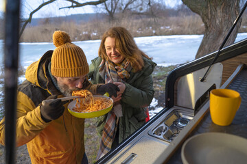 Hungry man eat lunch pasta from pan outdoors in wild, standing at mini camper back kitchen during winter journey vacation. Family spend time camping away in the wild together. Family journey concept. 