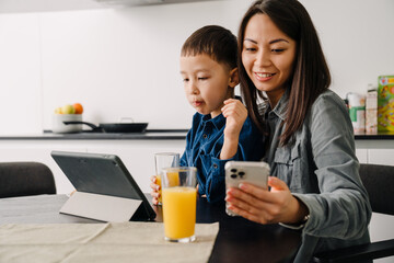 Happy asian woman using cellphone while having breakfast with her son