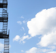 Silhouette Of A Fire Escape On A High-rise Building Against A Blue Sky With Clouds. Some Of The Stairs Are Broken. There Is Free Space For Text