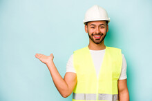 Young Laborer Hispanic Man Isolated On Blue Background Showing A Copy Space On A Palm And Holding Another Hand On Waist.