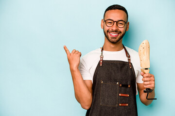 Wall Mural - Young hispanic shoemaker man isolated on blue background smiling and pointing aside, showing something at blank space.