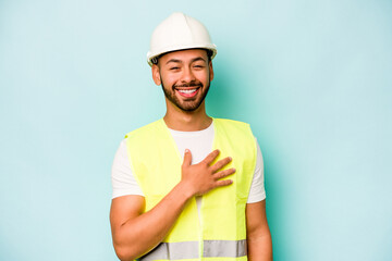 Wall Mural - Young laborer hispanic man isolated on blue background laughs out loudly keeping hand on chest.