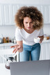 © LIGHTFIELD STUDIOS - curly woman with cup of coffee pointing with hand during video call on laptop in kitchen.