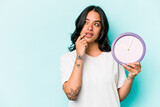 Fototapeta Na ścianę - Young hispanic woman holding a clock isolated on blue background relaxed thinking about something looking at a copy space.