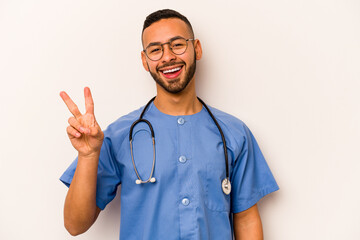 Wall Mural - Young hispanic nurse man isolated on white background joyful and carefree showing a peace symbol with fingers.