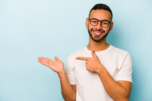 Young Hispanic Man Isolated On Blue Background Excited Holding A Copy Space On Palm.